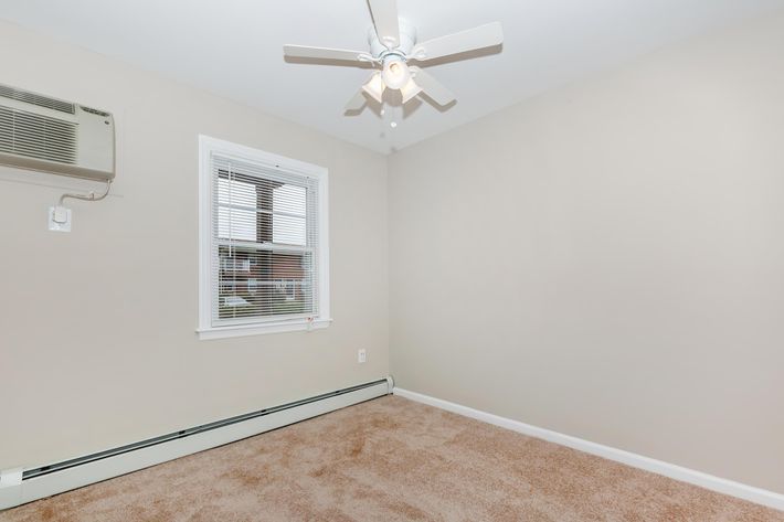 A small, empty room with beige walls and light brown carpet. There's a window with white blinds and an air conditioning unit mounted above it. A ceiling fan with white blades is installed in the center of the room. The space is well-lit and appears tidy.