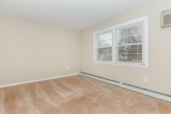 Bright room with light beige walls and a large window featuring white blinds. The floor is covered in light-colored carpet. There’s a baseboard heater along one wall, and minimal decoration creates a clean and empty space, suitable for various uses. Natural light fills the room.
