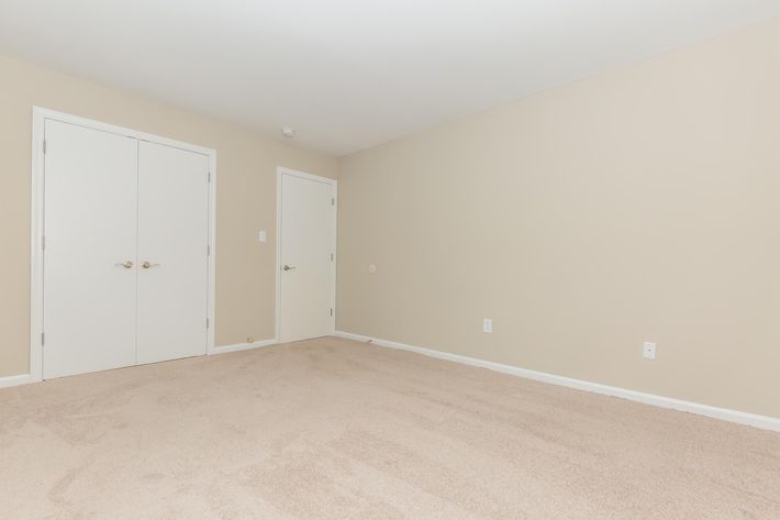 A spacious, empty room with beige walls and light carpet. There are two white closet doors visible on one wall and a simple light switch on another. The room is well-lit, conveying a clean and simple aesthetic, perfect for personalization.