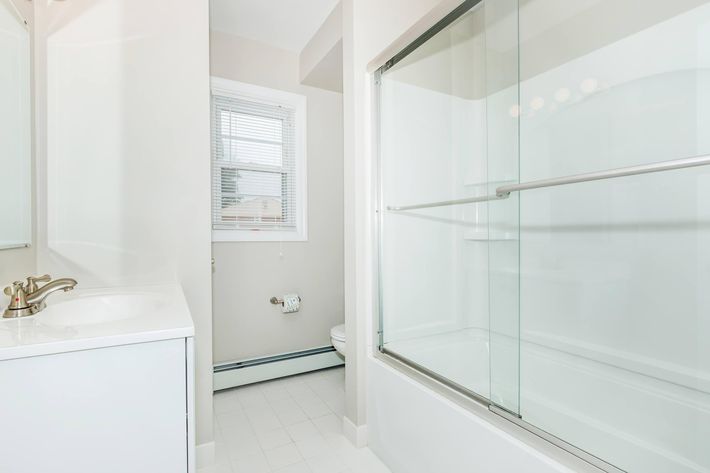 A modern bathroom featuring a bathtub with a glass sliding door, a white vanity with a sink, and a window allowing natural light. The walls are painted in a light color, and the floor is tiled in white. A small mirror is above the sink, and there is a towel holder beside the toilet.