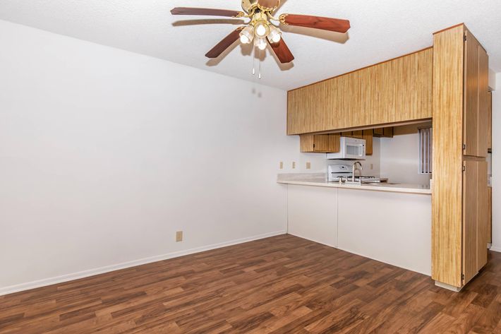 A spacious kitchen with a light wood finish, featuring a ceiling fan with lights. The kitchen includes a countertop, white appliances, and an open layout leading to a living area with laminate wood flooring. The walls are painted white, creating a bright and airy atmosphere.