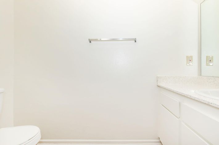 A clean, minimalist bathroom featuring a white wall, a toilet on the left, and a counter with a sink on the right. The counter has a light-colored granite surface, and there is a small mirror above it. A towel rack is mounted on the wall but is empty, emphasizing the spaciousness of the room.