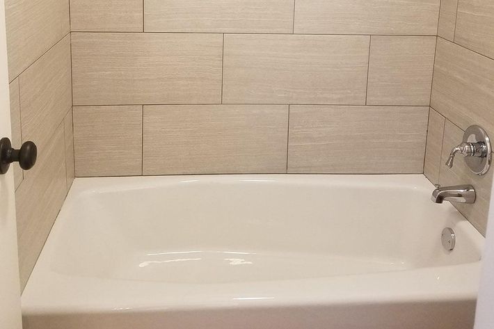 A clean, modern bathroom featuring a white bathtub set against light gray tiled walls. The tub has a sleek chrome faucet and a simple design, creating a minimalist look. The space is well-lit, enhancing the contemporary feel of the room.