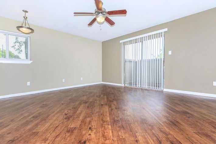 Spacious interior of a living room featuring hardwood-style flooring, a ceiling fan with wooden blades, and a sliding glass door with vertical blinds. The walls are painted in a soft beige, and there is a window beside the door allowing natural light to enter.