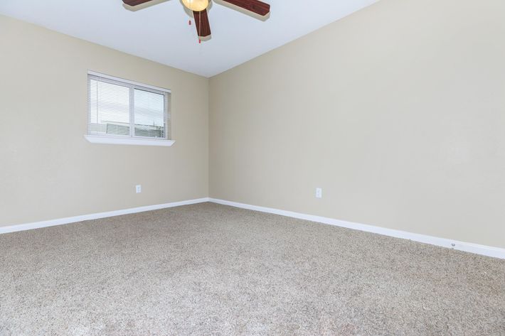 A spacious, empty room with beige walls and light brown carpet. It features a ceiling fan with wooden blades and a window with horizontal blinds, allowing natural light to enter. The room has no furniture and appears ready for decorating or moving in.
