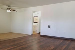 A spacious room featuring light-colored walls and a ceiling fan. The floor is a combination of tiles and hardwood. In the background, there's a doorway leading to a bathroom with visible fixtures. The overall ambiance is bright and inviting.