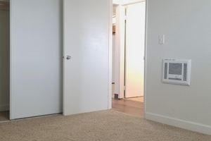 Interior view of a light-colored room featuring two doors, one leading to a closet and the other to a hallway. The floor is covered in beige carpet, and a wall-mounted heating unit is visible on the right. Natural light enters from the hallway, enhancing the spacious feel of the area.