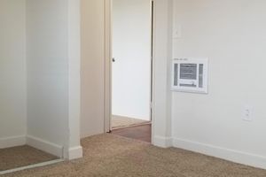 A view of a small, carpeted room with a doorway leading to another room. The walls are painted white, and there is a wall-mounted heater on the right. The floor is covered with light-colored carpet, and the doorway beyond is partially visible, suggesting a simple, clean interior space.