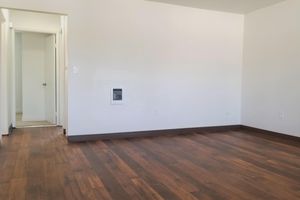 A vacant room featuring hardwood flooring and white walls. There is a doorway on the left leading to another space, and a wall-mounted heating unit on the right. The room is well-lit and free of furniture, creating an open and airy atmosphere.