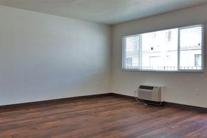 A bright, empty living room featuring light-colored walls and a large window with blinds. The floor is made of dark wood laminate, and there is a wall-mounted air conditioning unit in one corner. The space is uncluttered, creating a clean and minimalistic atmosphere.