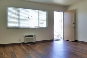 A bright, empty room with wooden flooring, featuring a window with blinds that lets in natural light. There's a wall-mounted air conditioning unit on the left, and a metal door on the right leading outside, partially open, with a grid pattern. The walls are painted a light color, creating a spacious feel.