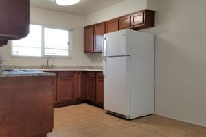 A clean, modern kitchen featuring light brown wooden cabinets and a white refrigerator. The space includes a sink under a window, with countertops made of light-colored stone. The floor is tiled in a neutral shade, and there is ample natural light entering the room.