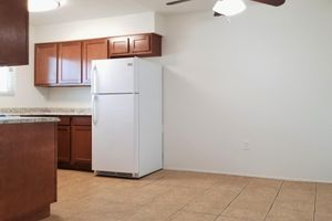 A clean, modern kitchen featuring wooden cabinets, a white refrigerator, and a tiled floor. The kitchen has a light-colored wall and a ceiling fan, with a spacious layout and a countertop that contrasts with the cabinetry. Natural light is coming from a window, creating a warm atmosphere.