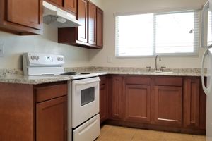 A modern kitchen featuring wooden cabinets, a white stove, and a refrigerator. Granite countertops complement the space, with a sink underneath a window allowing natural light to illuminate the room. The kitchen has a clean and organized appearance with neutral-colored tiles on the floor.