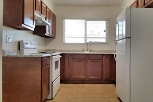 A modern kitchen featuring wooden cabinets, a countertop with a subtle granite pattern, a white stove and oven, a white refrigerator, and a window allowing natural light to enter. The floor has beige tiles, and the overall design is clean and functional.