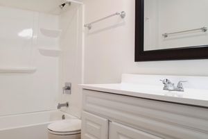 A clean and simple bathroom featuring a white bathtub with a shower, a white vanity with a sink, and a large mirror framed in black. The walls are painted a light color, and there are towel bars on either side of the mirror. The overall design is minimalistic and well-lit.