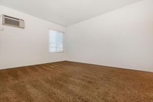 Empty room with light-colored walls and a large window covered by horizontal blinds. The floor is covered in brown carpet, and an air conditioning unit is mounted on the wall. The space appears clean and bright, suggesting it could be used for various purposes.