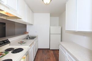 A clean, modern kitchen featuring white cabinets, a two-basin stainless steel sink, a stove with a vent, and a white refrigerator. The countertops are light-colored with a smooth finish, and the flooring is a warm wood tone. The walls are painted in a neutral shade, creating a bright and inviting atmosphere.