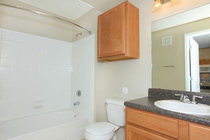 A clean and well-lit bathroom featuring a bathtub with a showerhead, a modern toilet, and a granite countertop with a sink. Light wood cabinetry is mounted above the toilet and next to the mirror, which reflects the room. The walls are painted in a light color, giving the space an airy feel.