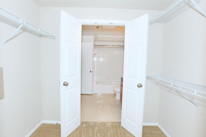 A view of a bathroom from an adjoining closet with two open white doors. The bathroom features a bathtub, toilet, and beige walls, while the closet has wire shelving on both sides. The floor is carpeted in a neutral color.