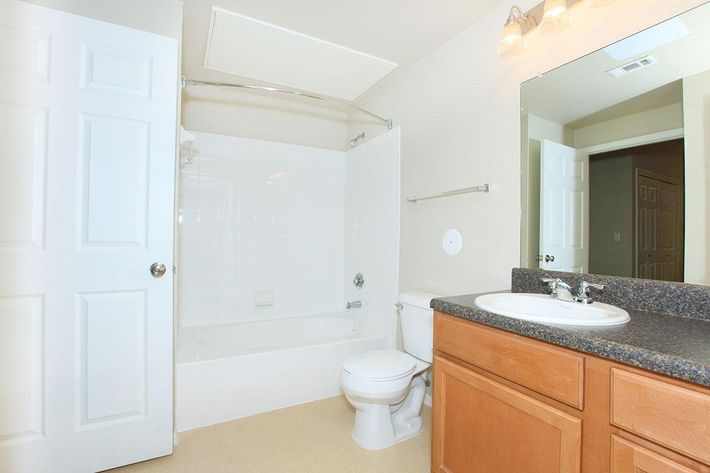 A clean and simple bathroom featuring a white bathtub with a shower, a toilet, and a double sink vanity with wooden cabinets. The walls are painted in neutral tones, and there's a large mirror above the sink. Soft lighting fixtures and a door leading to another room are visible.