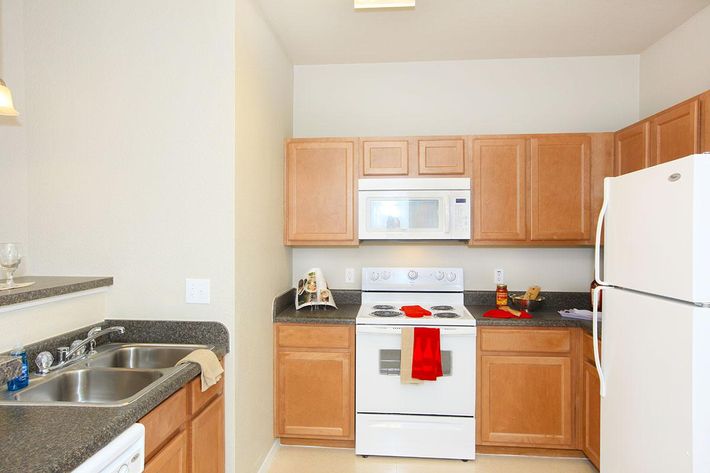 A modern kitchen featuring light wood cabinetry, a white refrigerator, an electric stove with an oven, a microwave above, and a double sink. Countertops are dark and uncluttered, with decorative items on the countertop and a red towel hanging near the stove.