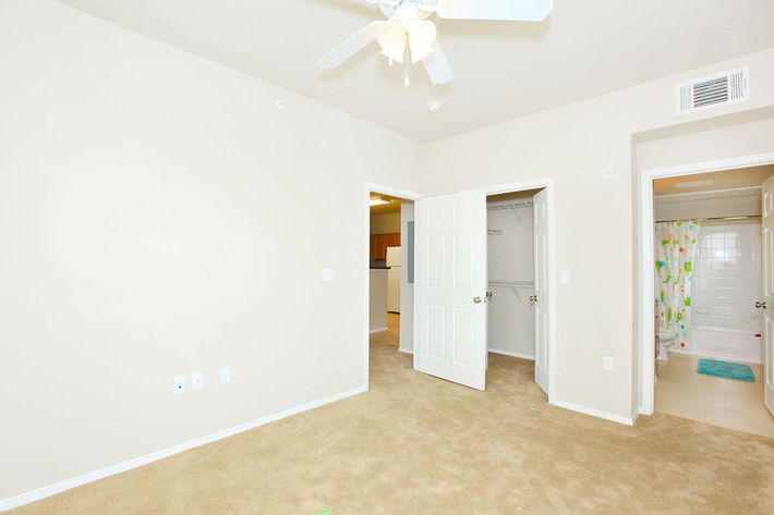 A light-colored room with beige carpet and plain walls, featuring two open doors. One door leads to a bathroom with a shower curtain. A ceiling fan is visible above, and there is a small section of the kitchen in the background, suggesting it's part of an apartment or small living space.