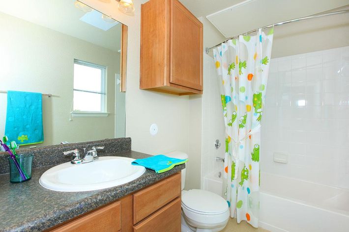 A bright and tidy bathroom featuring a brown wooden cabinet, a white sink, and a bathtub with a colorful shower curtain decorated with frogs. There’s a turquoise towel hanging on the wall and a blue washcloth on the countertop. Natural light is coming through a nearby window.