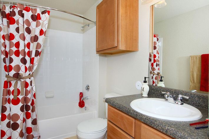 A clean bathroom featuring a white bathtub with a red circular-patterned shower curtain. The countertops are dark granite, and there's a white sink with modern fixtures. Wooden cabinets are above the sink, and a mirror reflects the space. A red towel hangs nearby, adding a pop of color.