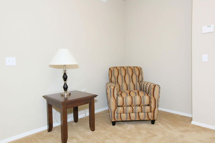A cozy corner featuring a patterned armchair with an oval design in warm colors, accompanied by a small wooden side table with a black lamp. The wall is painted a light beige, and the floor is covered with plush carpeting.