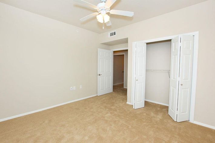 A light beige bedroom with light carpeting, featuring a ceiling fan. The room has two open white doors leading to a closet and another room, with plain walls and no furnishings, creating a spacious and empty atmosphere.