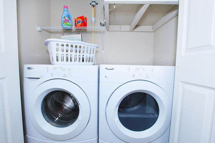 A laundry room featuring a stacked washer and dryer, a laundry basket, detergent bottles, and a shelving unit. The walls are painted in a neutral tone, and the space is organized and tidy.
