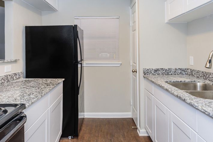 A modern kitchen featuring black appliances, including a refrigerator and stove. The countertops are made of gray speckled granite, and there are white cabinets above and below. A window with blinds allows natural light in, illuminating the space with light-colored walls and dark hardwood flooring.