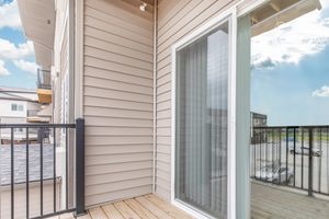 A corner balcony of a modern apartment, featuring a wooden deck and a sliding glass door. The exterior walls are clad in light beige siding, with a black metal railing enclosing the balcony. The view includes a parking area and blue sky with clouds in the background.