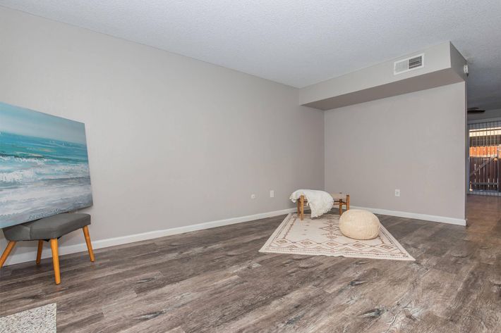 A minimalist living space featuring light gray walls, a wooden floor, and a large area rug. There is a small wooden stool with a beach-themed painting, a decorative pouf, and a simple wooden bench. The room has an open feel, with natural light coming from an entrance leading to an outdoor area.