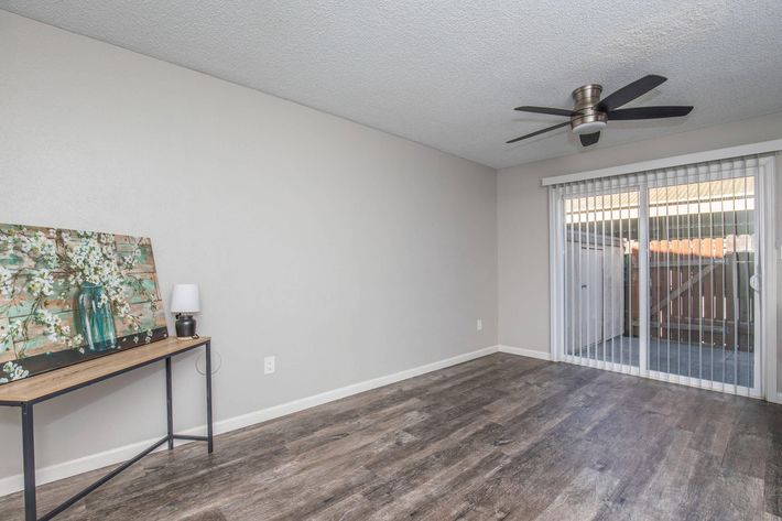 Spacious living area featuring a light-colored wall, modern ceiling fan, and laminate flooring. A simple desk with a decorative painting and lamp is positioned against one wall, while sliding glass doors lead to an outdoor space, allowing natural light to fill the room.
