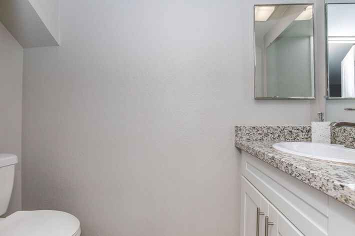 A clean, modern bathroom featuring a white toilet, a granite countertop with a sink, and a mirror above it. The walls are painted in light gray, creating a spacious and bright atmosphere. The overall design is contemporary and minimalistic.