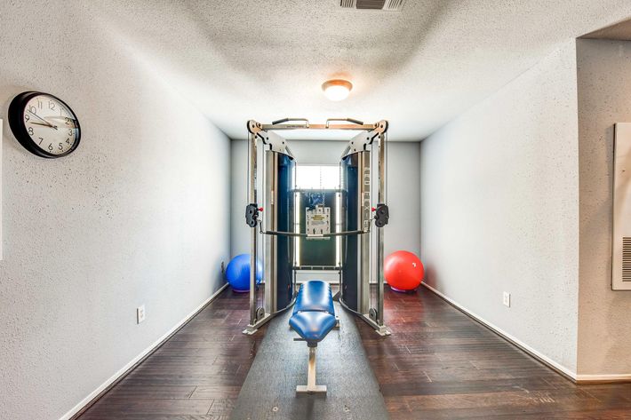 A small home gym featuring a multi-functional exercise machine in the center, a blue workout bench, and two large exercise balls (one blue and one red) on the floor. There is a clock on the wall and a window allowing natural light to enter the space. The walls are painted in a light color, and the flooring is dark wood.