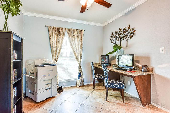 A home office featuring a desk with a computer and two chairs, a printer nearby, a window with curtains, and decorative wall art. The space has tiled flooring and a ceiling fan.