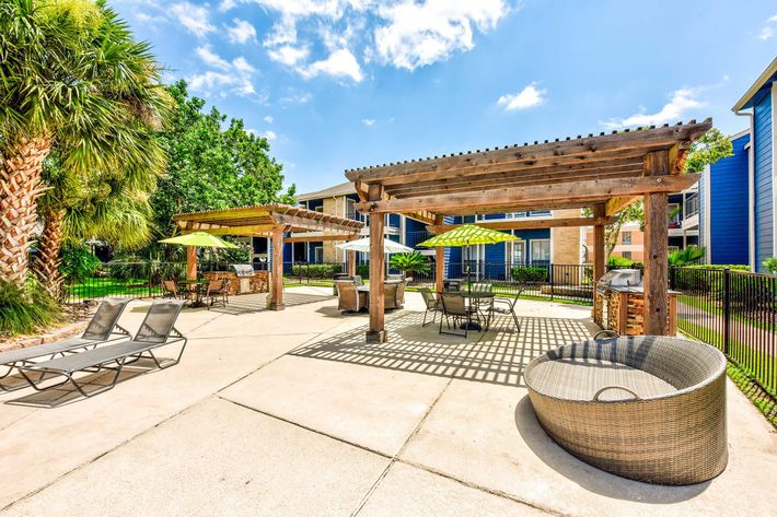 A sunny outdoor poolside area featuring lounge chairs, shaded seating under wooden pergolas, vibrant green umbrellas, and palm trees. The setting has a paved surface and includes a barbecue grill, surrounded by apartment buildings.