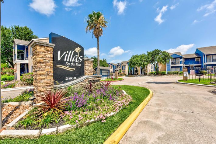 Sign featuring "Villas By the Bay," located at 2800, surrounded by landscaped gardens with palm trees and colorful flowers, leading to an entrance of apartment buildings under a clear blue sky.