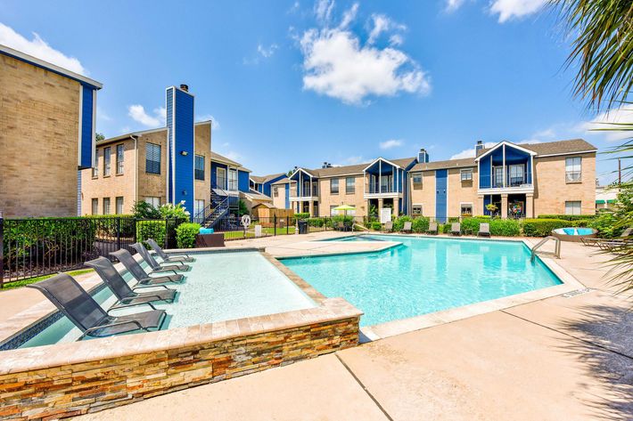 A sunny apartment community featuring a large swimming pool with lounge chairs around it, surrounded by well-maintained landscaping and residential buildings in the background.
