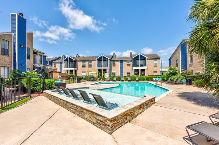 A swimming pool surrounded by lounge chairs, with apartment buildings in the background under a blue sky with scattered clouds.