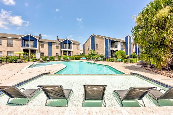 A serene view of a swimming pool surrounded by well-maintained lawns and residential buildings. Comfortable lounge chairs are positioned along the pool's edge, under a sunny blue sky with a few clouds.