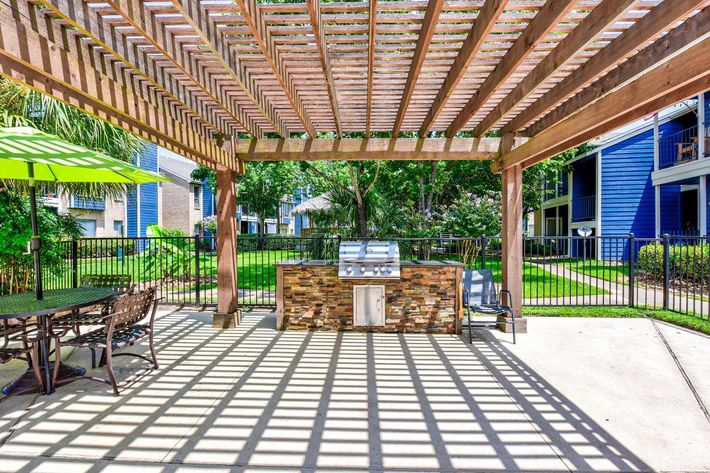 Outdoor patio space featuring a stone barbecue grill under a wooden pergola, with a round table and chairs nearby, surrounded by green grass and trees, and apartment buildings in the background.