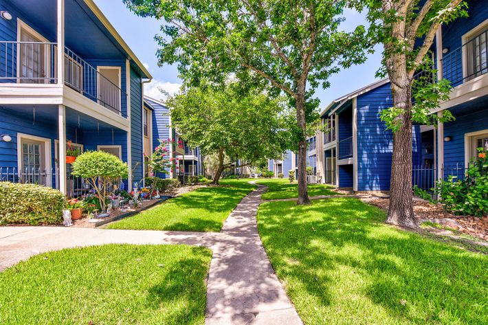A landscaped pathway winding through a residential complex with blue buildings on either side, surrounded by greenery and trees.