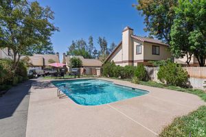 A well-maintained outdoor swimming pool surrounded by a concrete patio with lounge chairs and a table under a pink umbrella. Lush greenery and trees provide a tranquil setting, with residential buildings in the background against a clear blue sky.