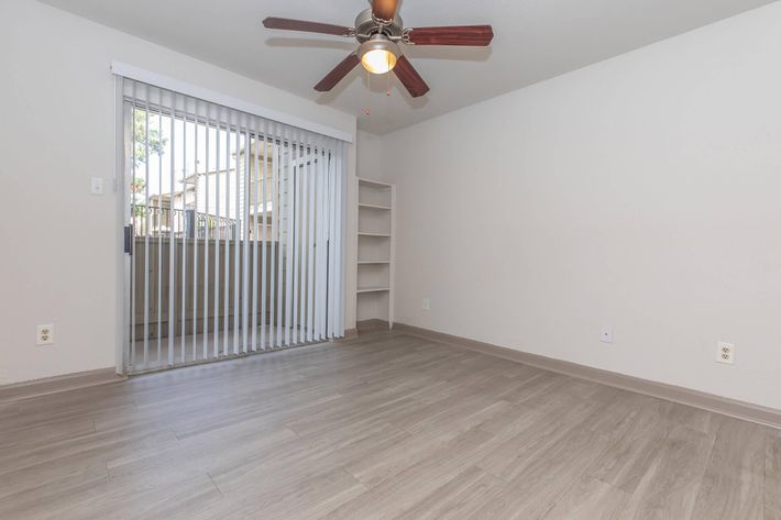Empty interior of a room featuring a ceiling fan, light-colored walls, and wood-like flooring. A set of vertical blinds covers a glass door that leads to a small balcony. There is a built-in shelf on one side, creating a minimalist and spacious appearance.