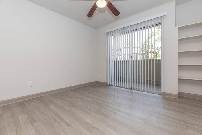 A spacious, empty room features light-colored laminate flooring and a ceiling fan. The walls are painted in a neutral tone, and there are vertical blinds covering a large window that opens to a balcony or outdoor area. A built-in shelf is present on one side of the room, enhancing the minimalist design.