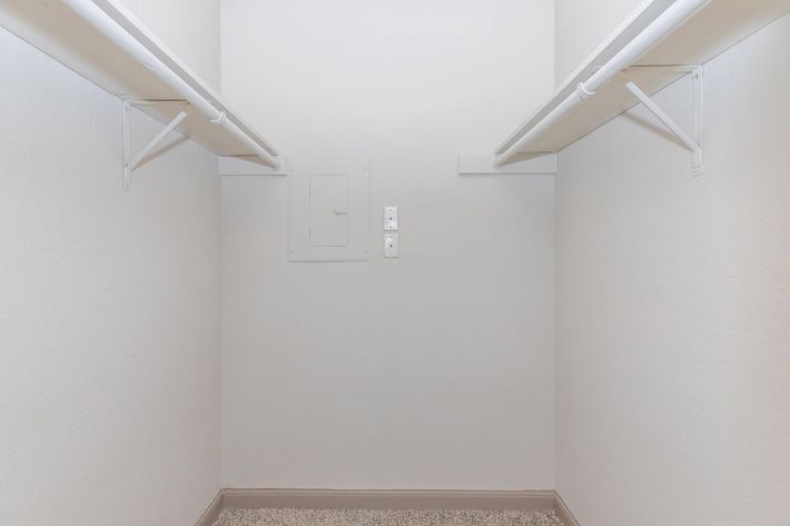 Empty closet with two white hanging rods on either side against light-colored walls. The floor is carpeted, and there is a small electrical panel on the back wall. The space appears clean and uncluttered, providing a blank canvas for storage.
